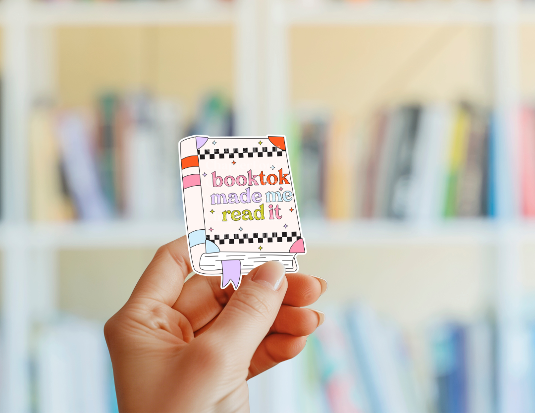 Hand holding a colorful book-shaped card with 'booktok made me read it' text against a blurred bookshelf background.