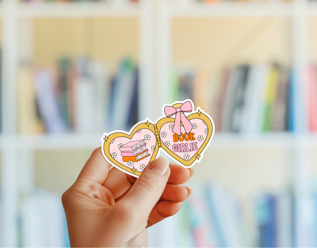 A double heart-shaped locket sticker with 'Book Girl' text on the right and 3 stacked books design on the left, hand holding in front of a blurred bookshelf.