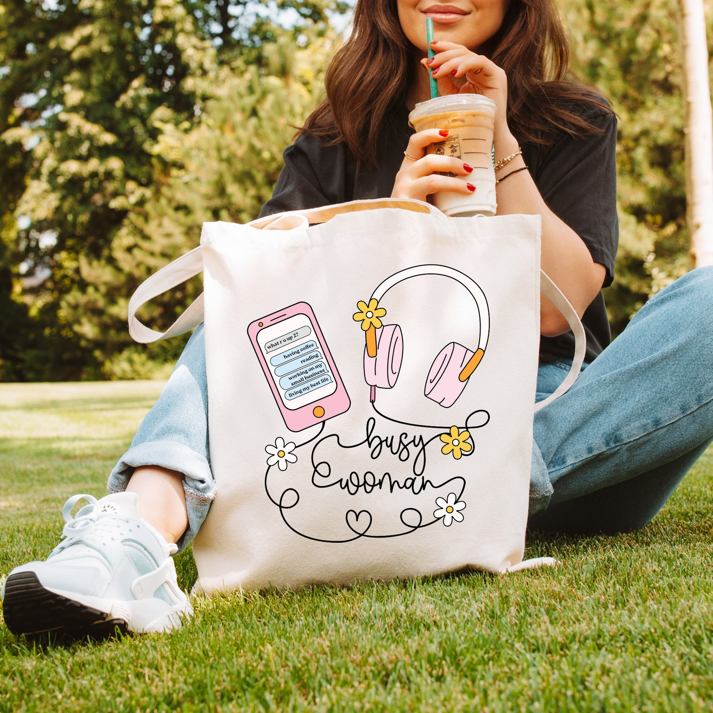 A person sitting on grass holding a tote bag with 'busy woman' text and illustrations of a phone, headphones, and flowers printed on it.