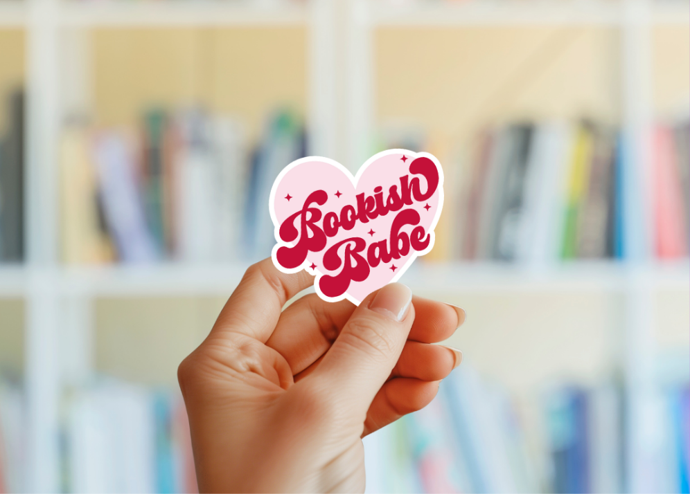 A hand holding a pink heart-shaped sticker with the red text 'Bookish Babe' on it, with blurred bookshelves in the background.