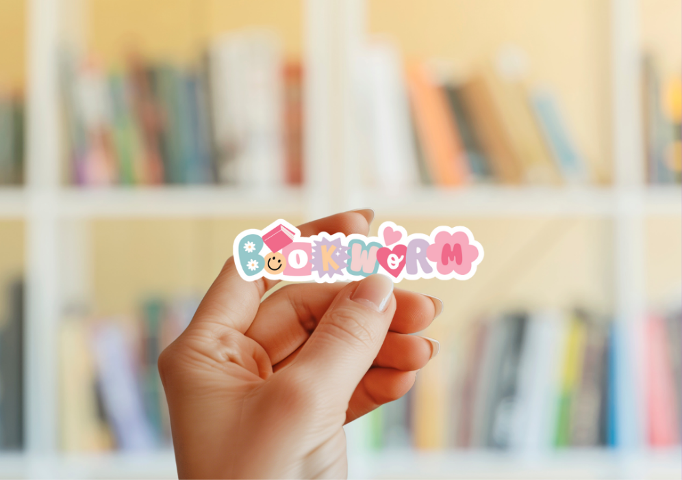 A hand holding a sticker with the word 'BOOKWORM' displayed against a blurred background of a bookshelf.