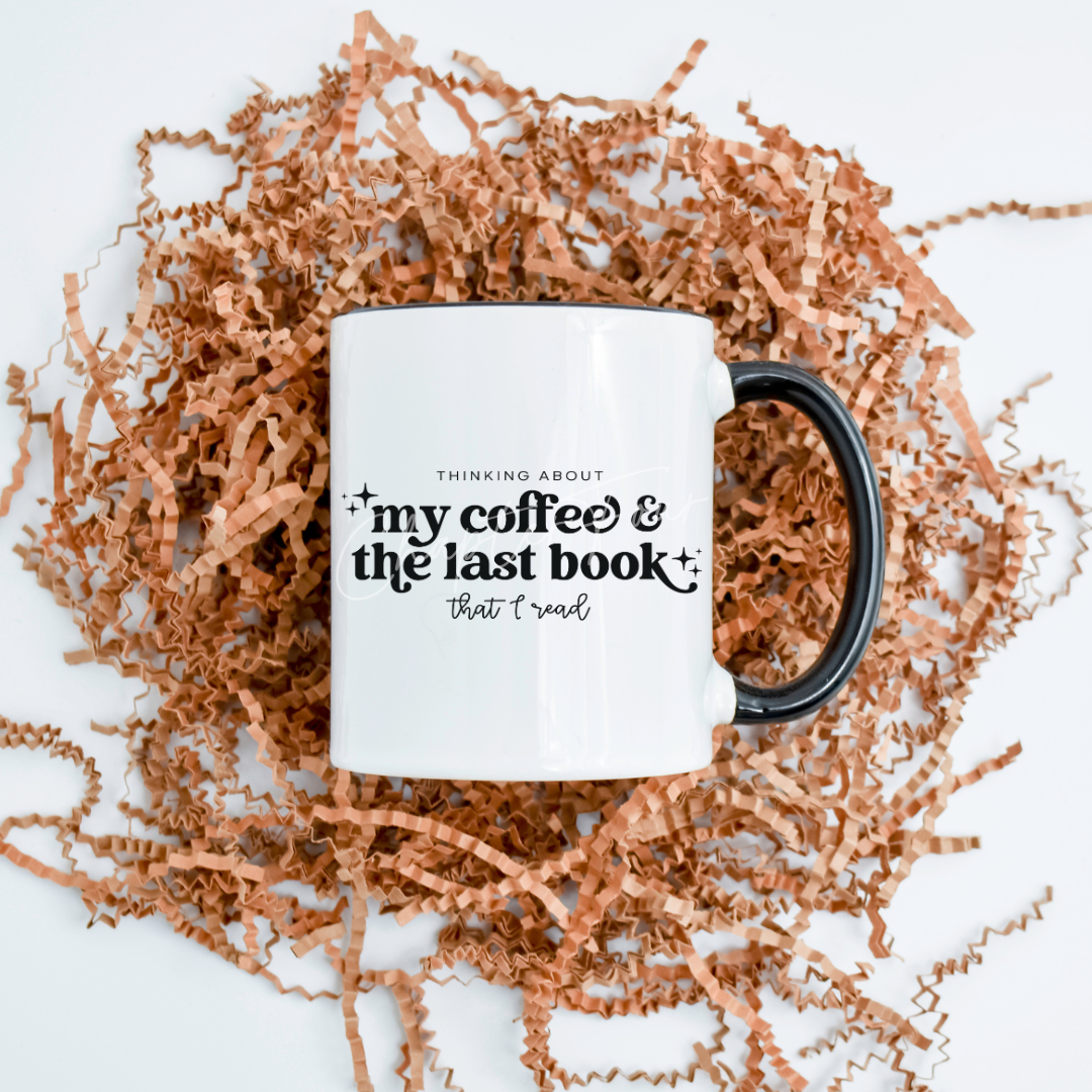 A white ceramic mug with black text and a black handle, featuring the phrase 'Thinking about my coffee & the last book that I read' printed on it, displayed on a bed of brown shredded filling.