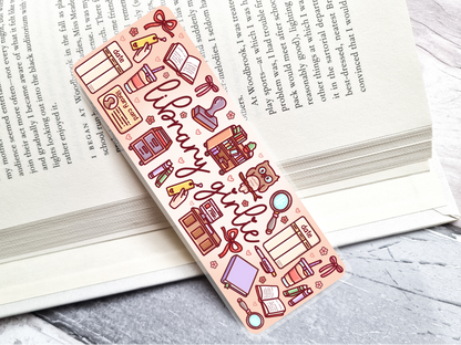Colorful bookmark with library-themed illustrations and 'library girlie' text, laying on an open book
