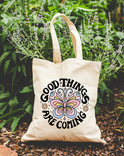 A beige cotton tote bag with the text 'GOOD THINGS ARE COMING' printed on it, displayed in a natural setting with plants in the background.