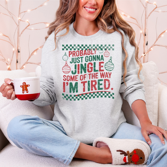 Person wearing a Christmas-themed sweatshirt that says Probably Just Gonna Jingle Some Of The Way I'm Tired and holding a mug, sitting on a couch.