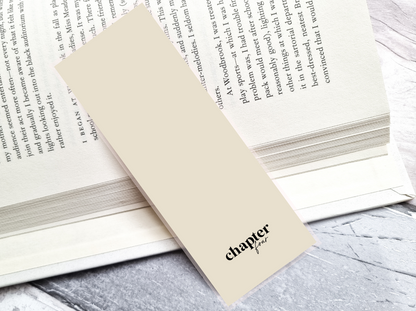 A beige paper bookmark with the black text 'chapter four' printed on it, resting on top of an open book with printed pages. The bookmark is photographed on a light gray stone-textured surface.