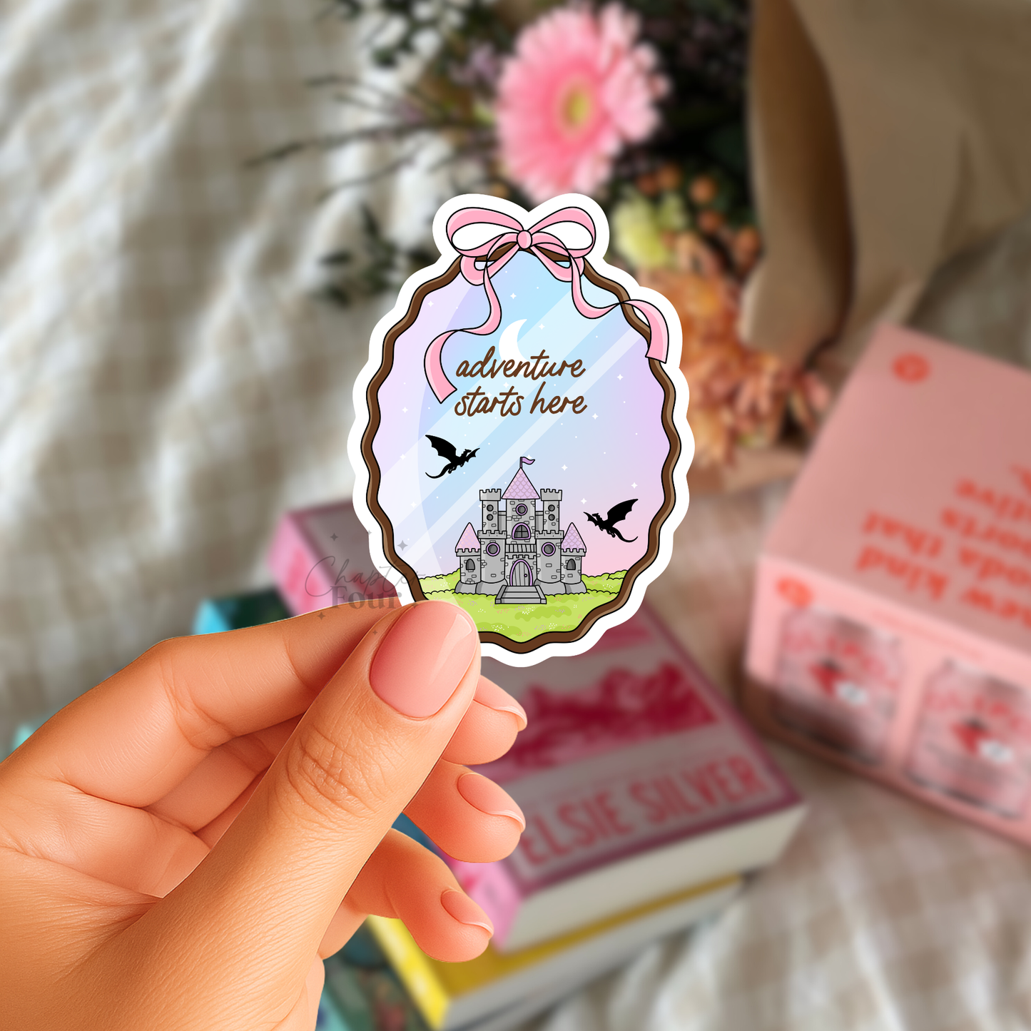 Hand holding a sticker with a castle design and 'adventure starts here' text, with books and flowers in the background.