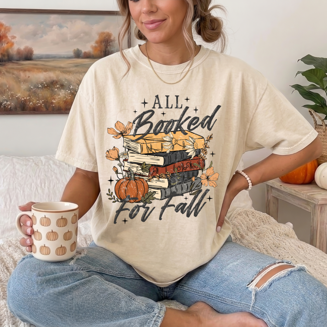 A woman sitting on a bed wearing a cream-colored graphic t-shirt that reads 'ALL Booked For Fall' with a stack of books, pumpkins, and autumn leaves. She is holding a mug with a pumpkin pattern.