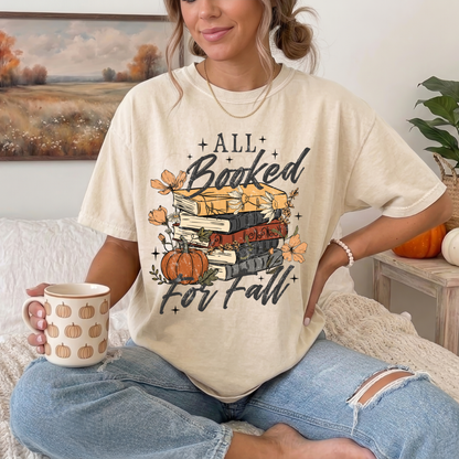 A woman sitting on a bed wearing a cream-colored graphic t-shirt that reads 'ALL Booked For Fall' with a stack of books, pumpkins, and autumn leaves. She is holding a mug with a pumpkin pattern.