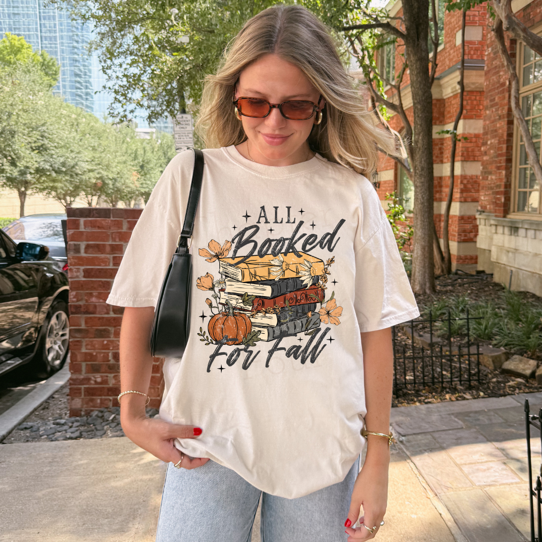 A woman wearing a cream-colored graphic t-shirt with a 'All Booked For Fall' autumn book stack design, standing outdoors on a sidewalk near a brick building and trees.