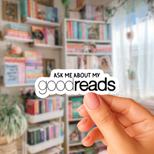 Hand holding a sticker with 'Ask me about my goodreads' text in front of bookshelves.