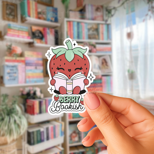 Hand holding a sticker with a strawberry character reading a book, labeled 'Berry Bookish', in front of a bookshelf.