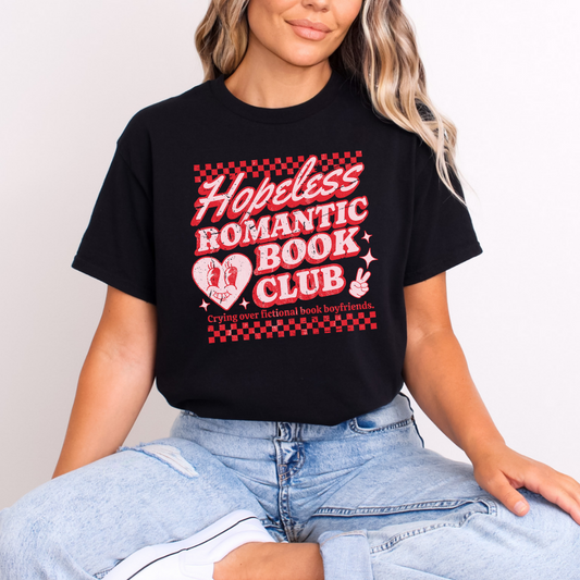 A person wearing a black t-shirt with 'Hopeless Romantic Book Club' text and heart graphics on it, sitting on a surface.