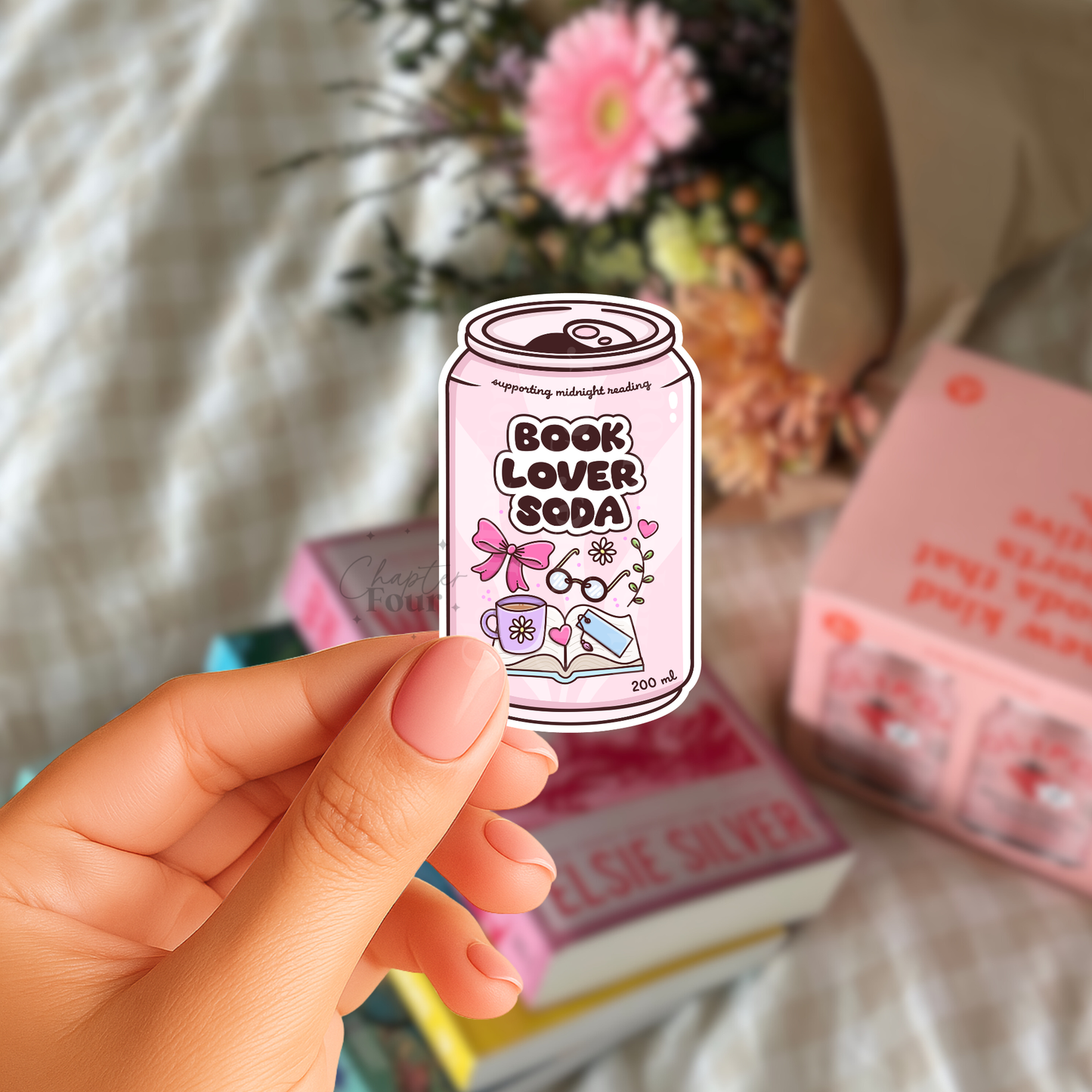 Hand holding a light pink 'Book Lover Soda' can-shaped vinyl sticker with books and flowers in the background