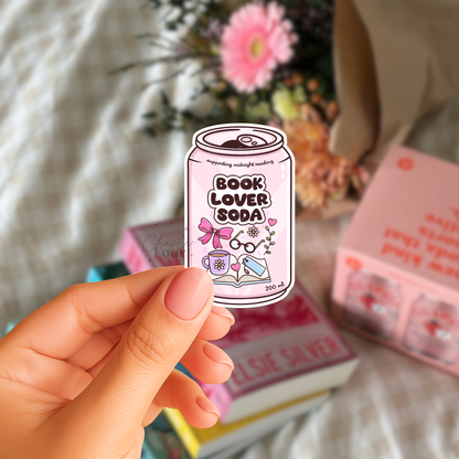Hand holding a light pink 'Book Lover Soda' can-shaped vinyl sticker with books and flowers in the background