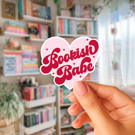 Pink heart-shaped sticker with 'Bookish Babe' red text held in front of a bookshelf.