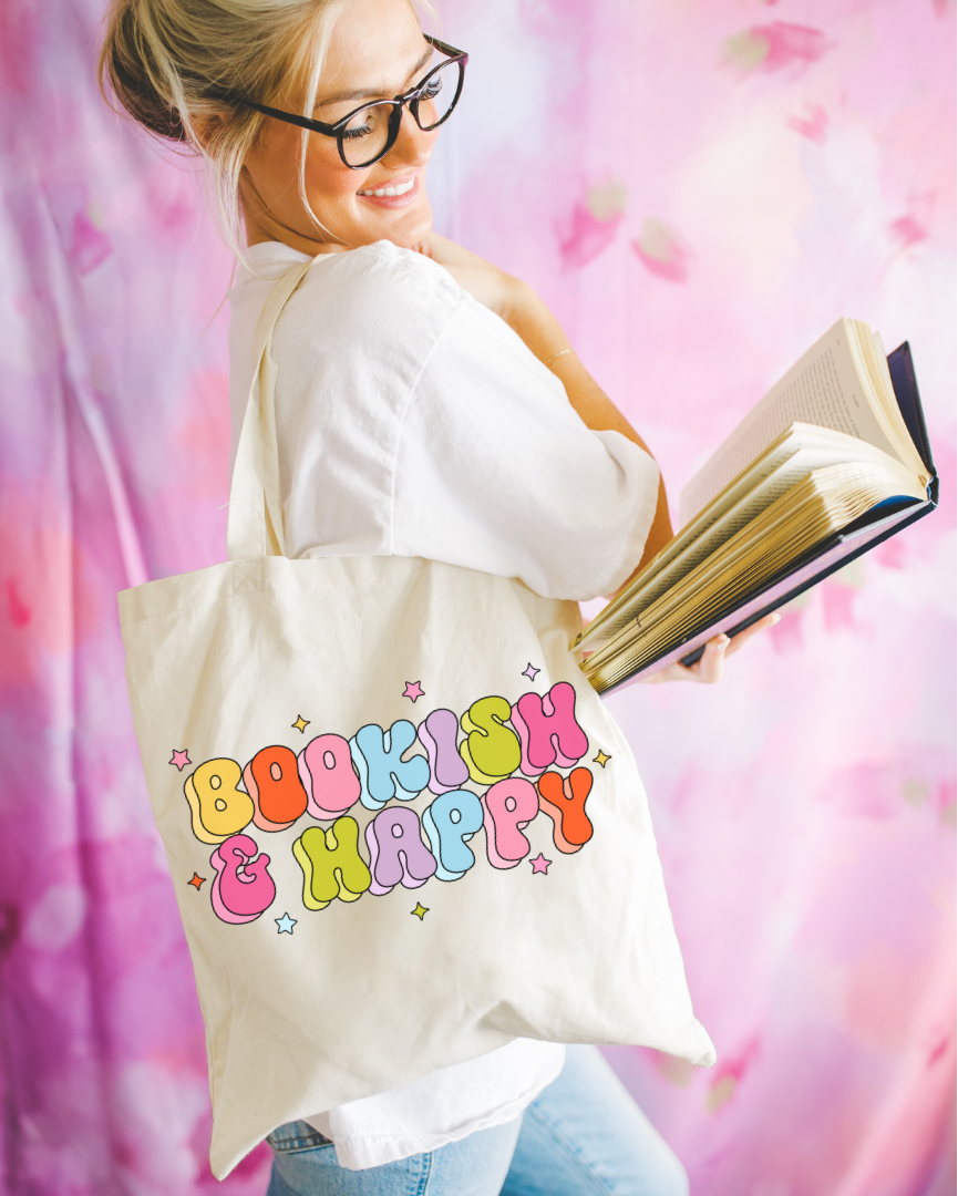A person holding an open book and wearing a white shirt with a tote bag that has 'BOOKISH & HAPPY' printed in colorful letters.