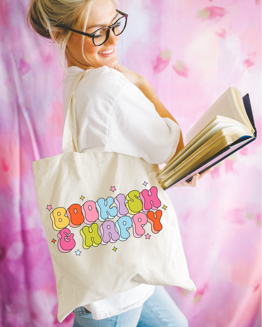 A person holding an open book and wearing a white shirt with a tote bag that has 'BOOKISH & HAPPY' printed in colorful letters.