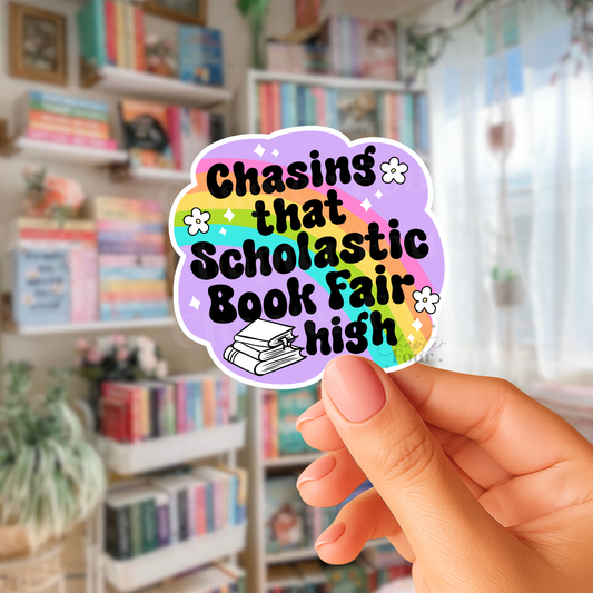 Hand holding a colorful sticker with 'Chasing that scholastic book fair high' text in a library setting