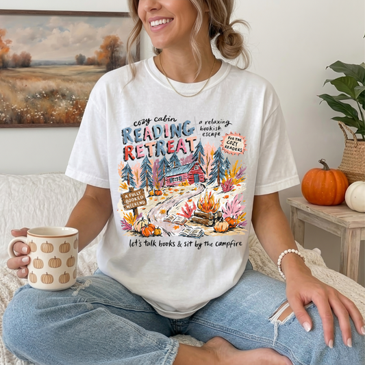 A woman sitting cross-legged indoors wearing a white graphic T-shirt with a colorful autumn cabin and reading retreat design, paired with light blue jeans. She holds a mug with pumpkin patterns, with a framed landscape, pumpkins, and a plant in the background.