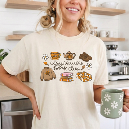A woman wearing a cream-colored graphic t-shirt with a cozy book club design, featuring illustrations of a sweater, mug, teapot, book, and cookies. She is holding a green mug with white flowers in a bright kitchen setting.