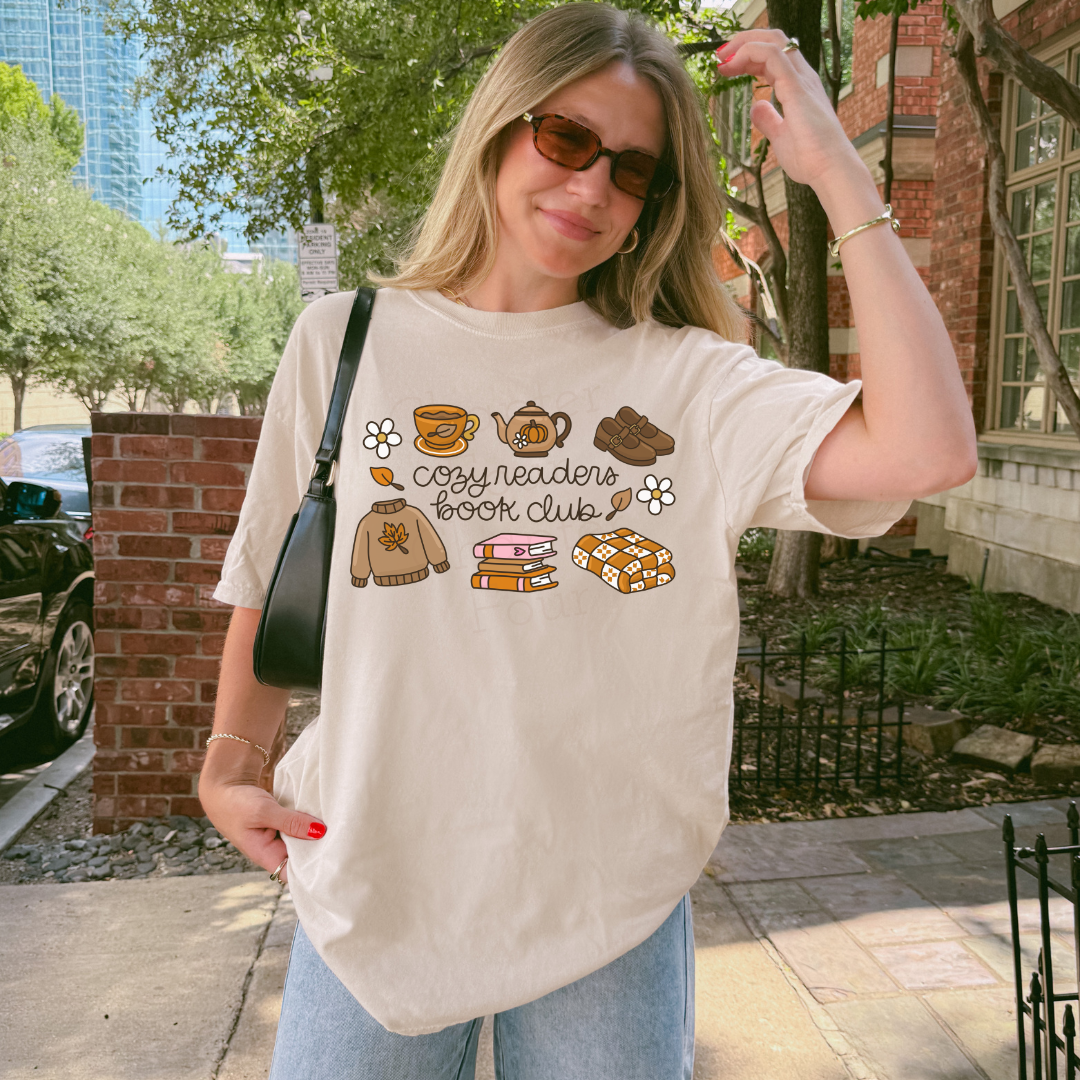 A woman wearing a cream-colored graphic t-shirt with a 'cozy readers book club' design featuring illustrations of a sweater, mug, teapot, book, and cookies. She is standing outdoors on a sidewalk next to a brick wall and trees.