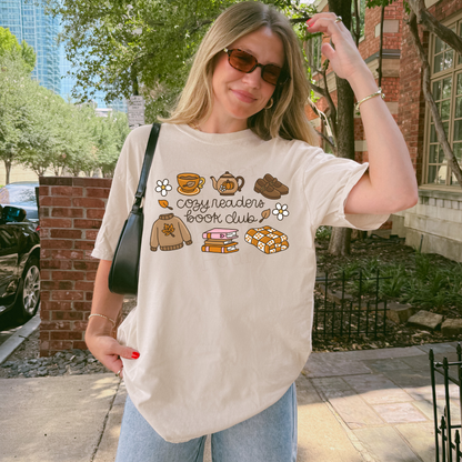 A woman wearing a cream-colored graphic t-shirt with a 'cozy readers book club' design featuring illustrations of a sweater, mug, teapot, book, and cookies. She is standing outdoors on a sidewalk next to a brick wall and trees.