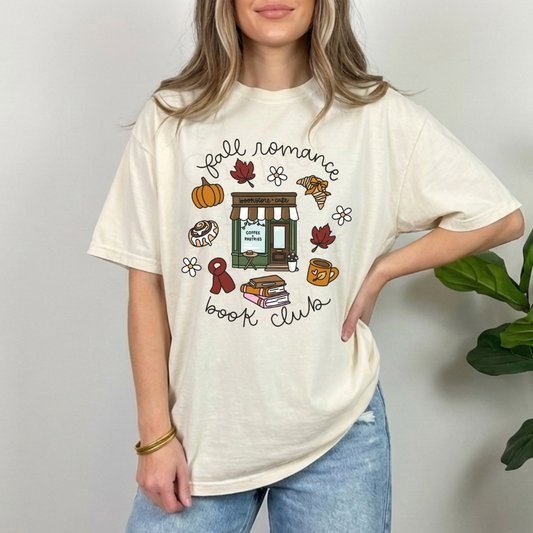 A woman wearing an oversized cream-colored graphic t-shirt with a 'Fall Romance Book Club' illustration featuring autumn leaves, a pumpkin, a book, and a coffee cup, paired with blue jeans.
