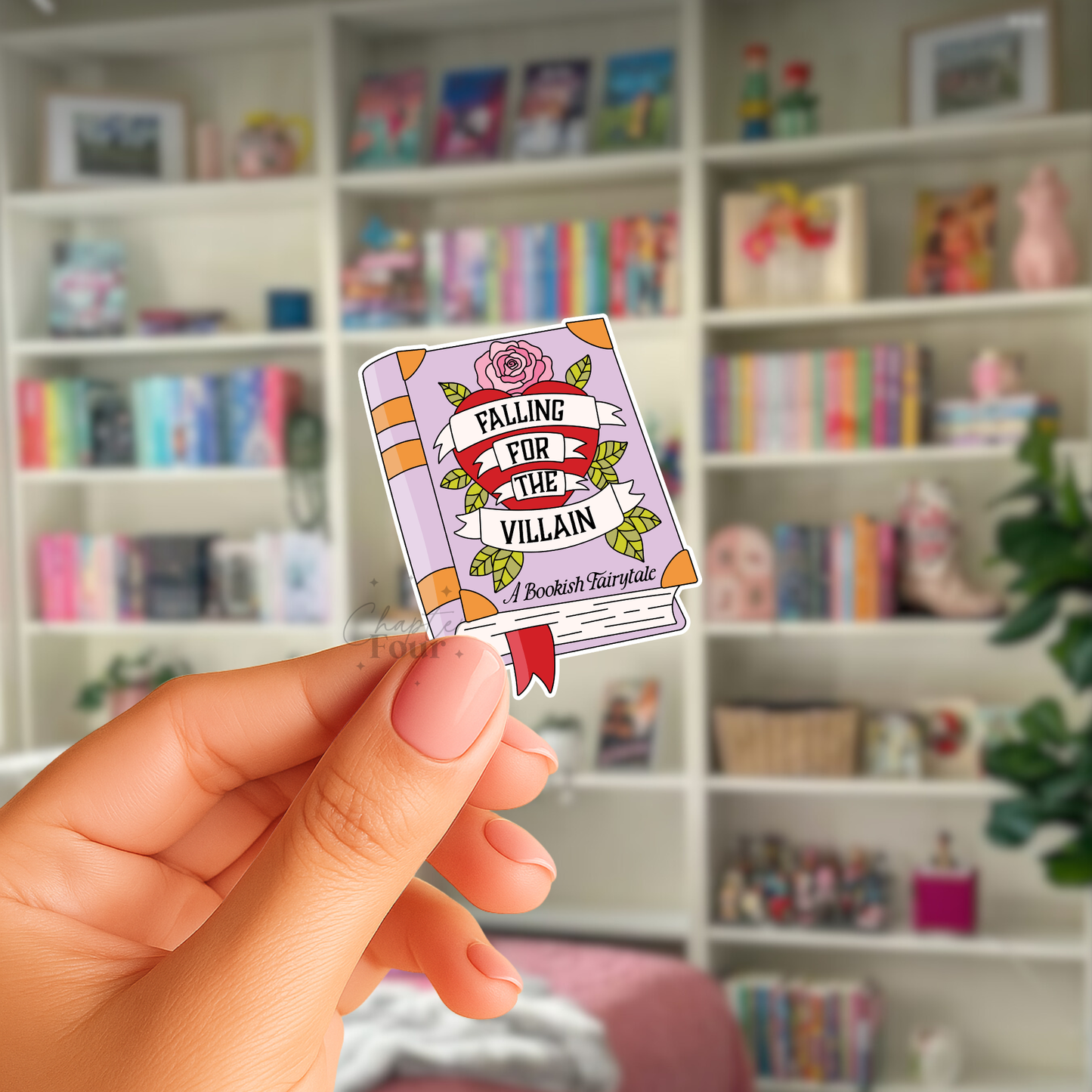 Hand holding a small book-shaped sticker with 'Falling for the Villain, A Bookish Fairytale' text in front of a bookshelf.