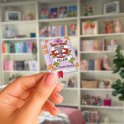 Hand holding a small book-shaped sticker with 'Falling for the Villain, A Bookish Fairytale' text in front of a bookshelf.