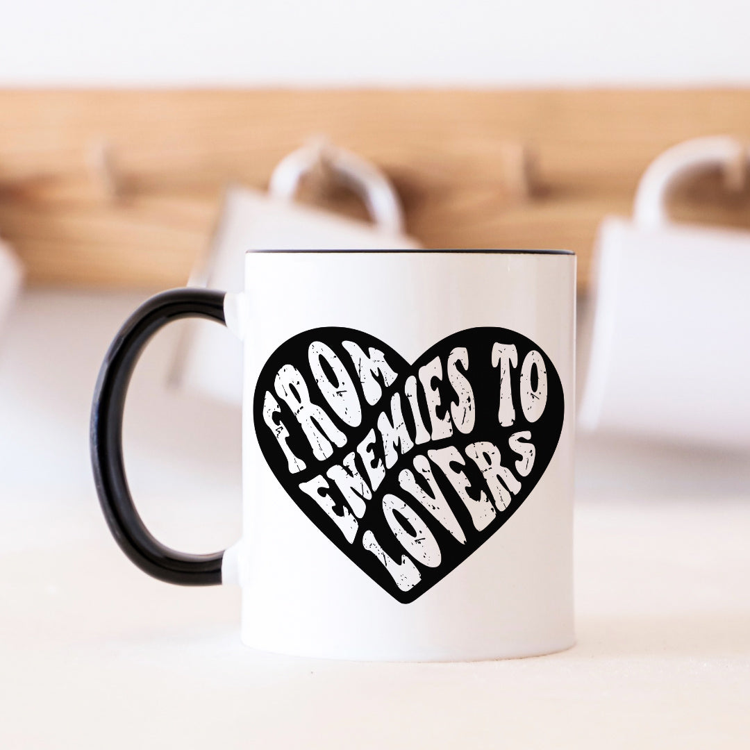 White mug with black handle and 'From Enemies to Lovers' text in a heart shape on a blurred background.