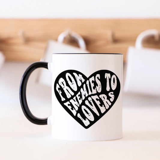 White mug with black handle and 'From Enemies to Lovers' text in a heart shape on a blurred background.