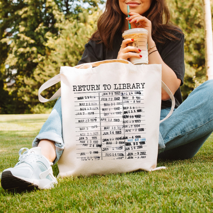 A person sitting on grass holding a tote bag with 'RETURN TO LIBRARY' printed on it, also holding a drink with a straw.