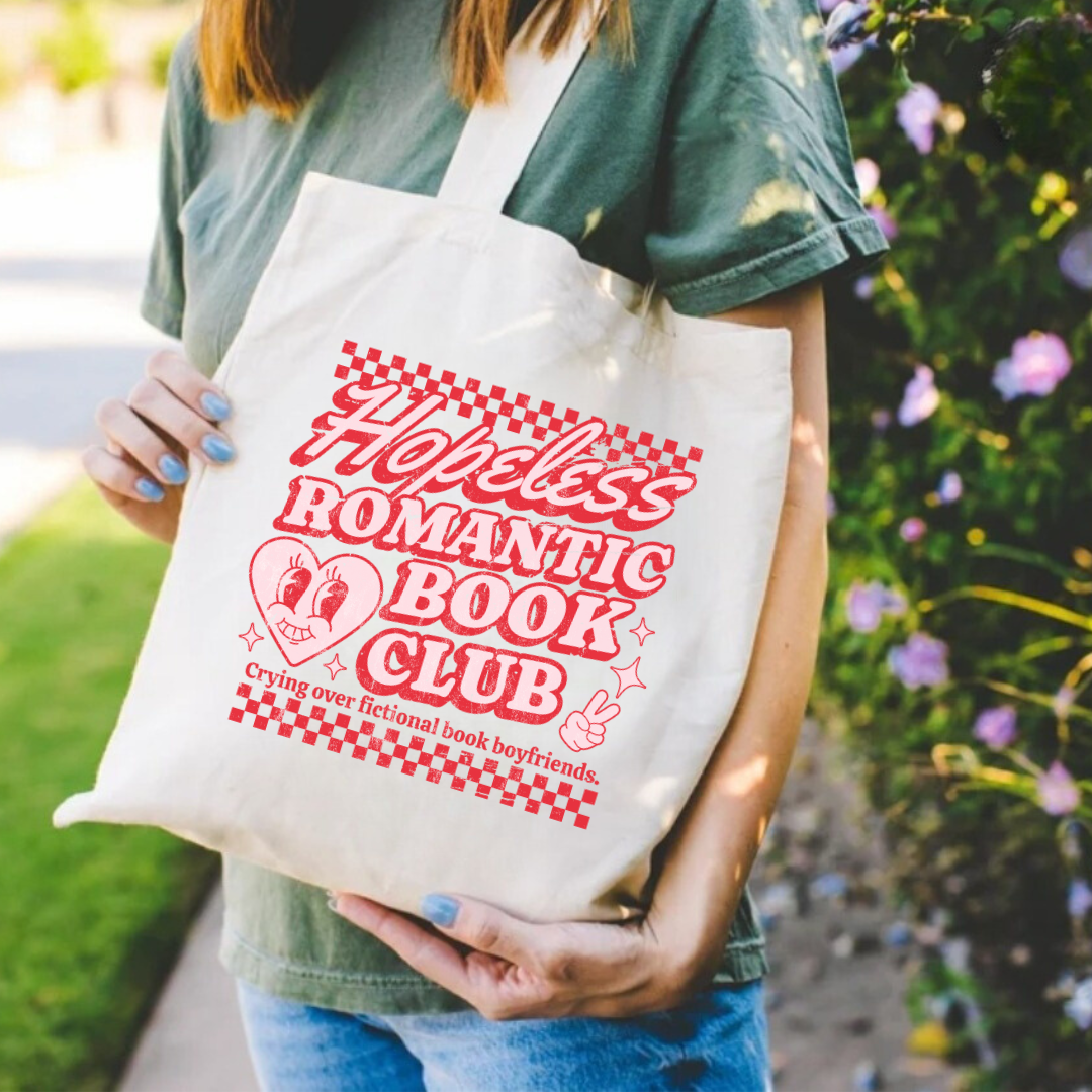 A person holding a beige cotton tote bag with the text 'Hopeless Romantic Book Club' printed on the front, along with a graphic of a heart and a book.