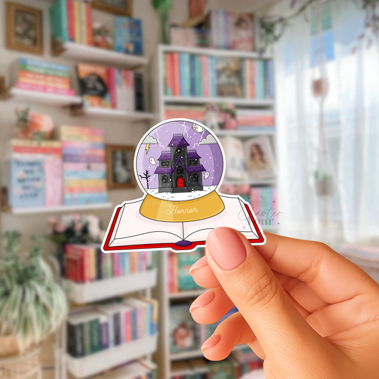 Hand holding a sticker of a purple and black haunted house in a globe with 'horror' text below, sitting on a book with a blurred bookshelf background