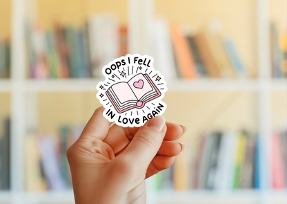 A hand holding a round sticker with a illustration of a book and the phrase 'Oops I Fell In Love Again' on it, with a blurred background of a bookshelf.