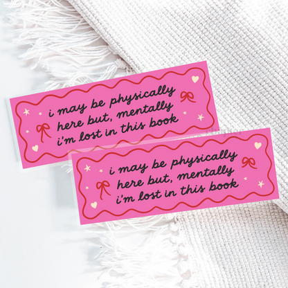 Two pink bookmarks with wavy red borders and black cursive text reading 'i may be physically here but, mentally i'm lost in this book', styled on a white textured blanket.