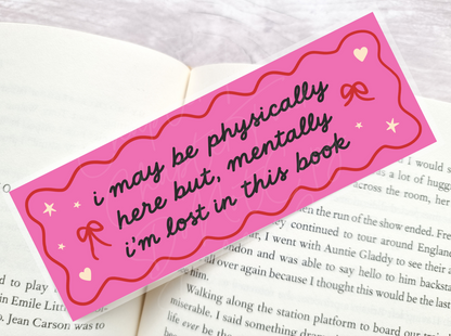 A pink bookmark with wavy edges lies across an open book. The bookmark has black script text that reads, 'i may be physically here but, mentally i'm lost in this book,' decorated with small red bows, white stars, and hearts.