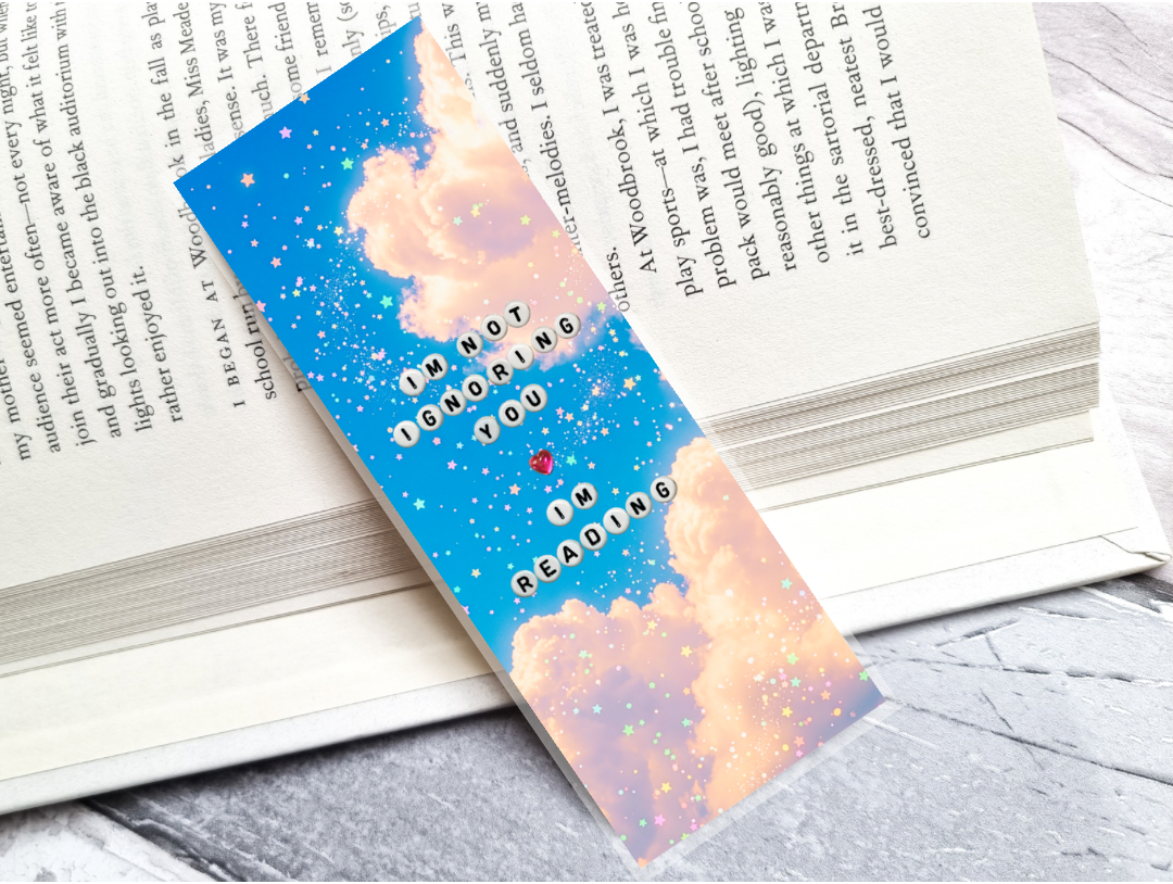 A decorative book bookmark with a blue and pink cloud design, glittery gold speckles, and the text 'I'M NOT IGNORING YOU IN READING' with a small red heart, shown resting on open books.