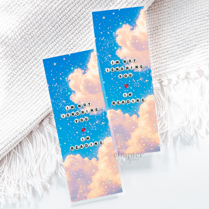 A decorative book bookmark with a blue and pink cloud design, glittery gold speckles, and the text 'I'M NOT IGNORING YOU IN READING' with a small red heart, placed on a white textured blanket with fringe.