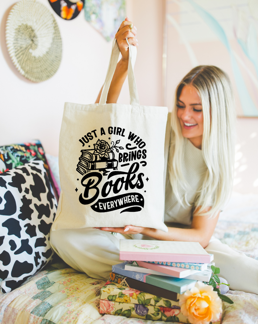 A person holding a beige tote bag with the text 'Just A Girl Who Brings Books Everywhere' printed on it, sitting beside a pile of books and decorative items.