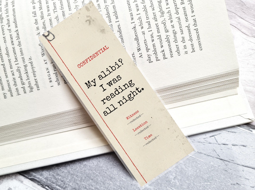 A cream-colored paper bookmark with black text reading 'My alibi? I was reading all night.' and red accents, resting on an open book over a gray marble surface.