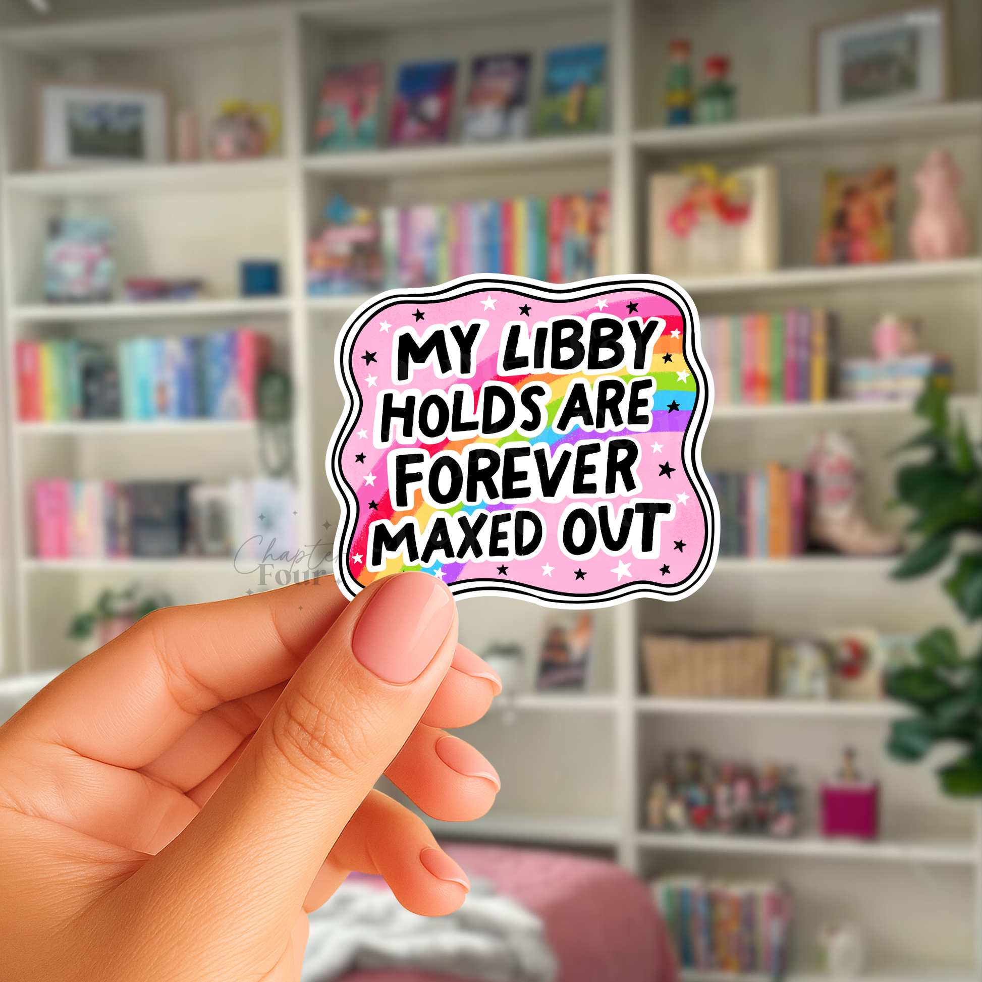 Hand holding a colorful sticker with 'My libby holds are forever maxed out' text in front of a bookshelf.