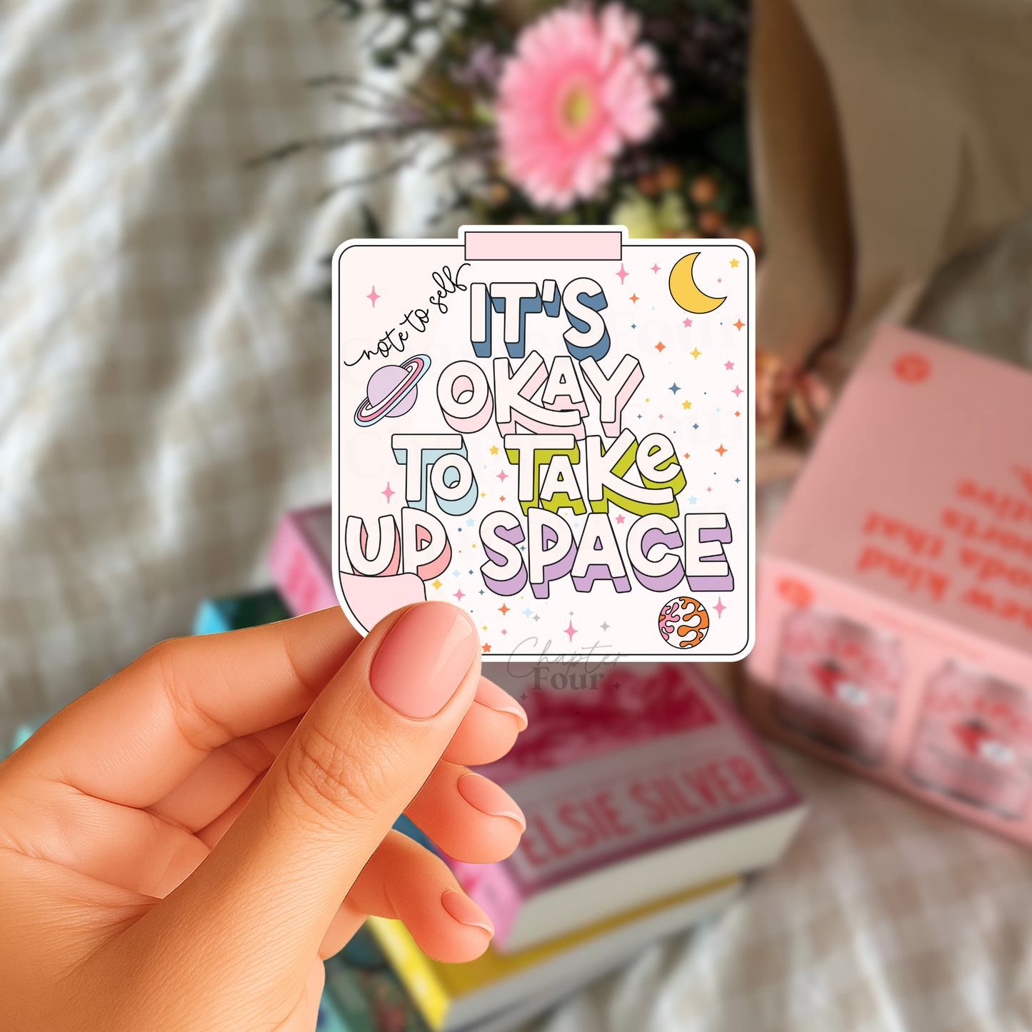 Hand holding a colorful sticker with 'note to self: It's okay to take up space' text, against a blurred background.