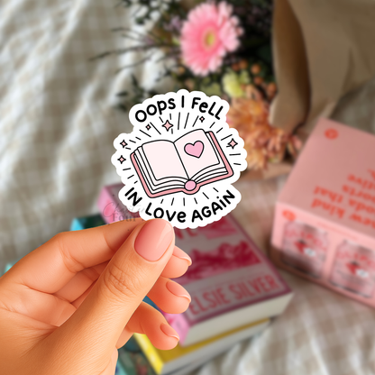 Hand holding a sticker with 'Oops I Fell in Love Again' text and book illustration, with books in the background.