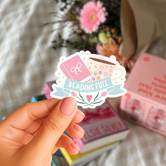 Hand holding a 'Reading Fuel' sticker with books and flowers in the background