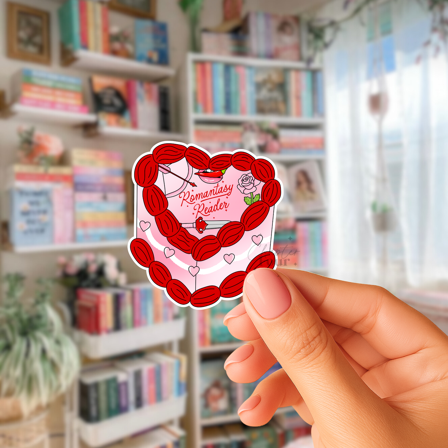 Red heart-shaped cake sticker with 'Romantasy Reader'  text held by a hand in front of a bookshelf.
