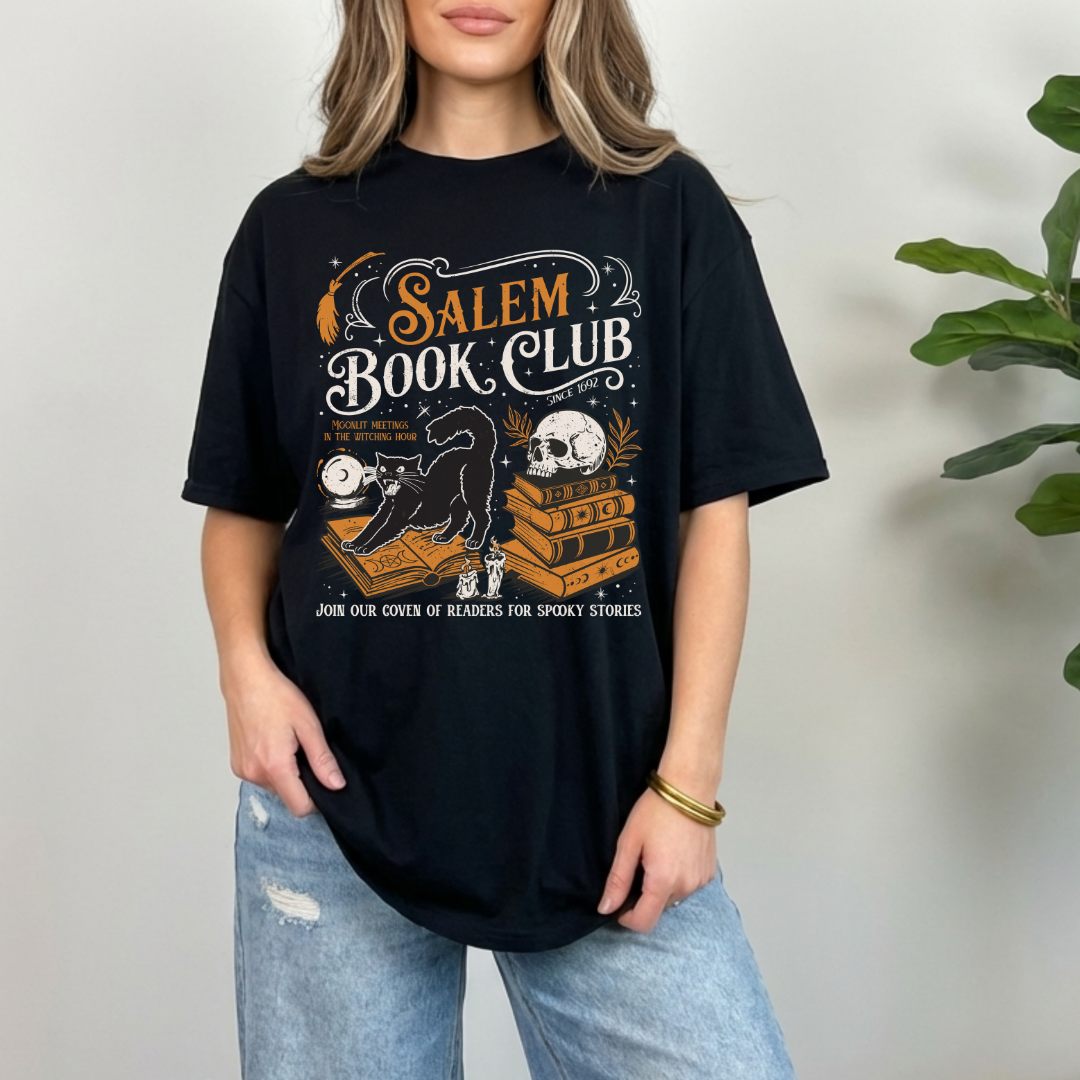 A woman wearing a black graphic t-shirt with a 'Salem Book Club' design, paired with light wash blue jeans, standing against a neutral wall with a green plant in the background.
