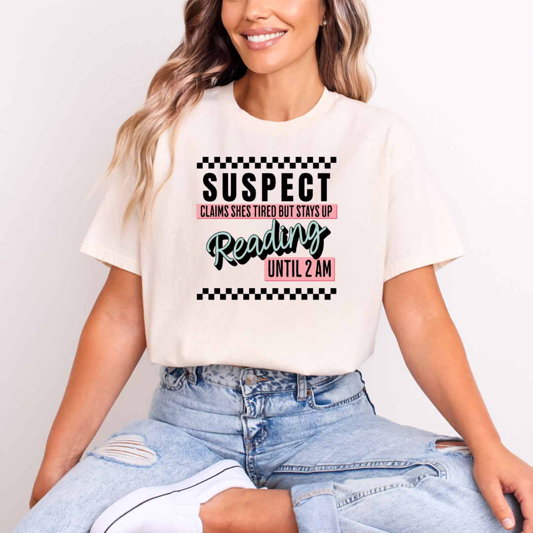 A person wearing a beige t-shirt with the text 'SUSPECT CLAIMS SHE'S TIRED BUT STAYS UP READING UNTIL 2 AM' printed on the front.