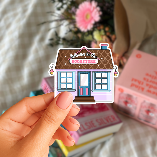 Hand holding a sticker of a bookstore with 'take me to the bookstore' text, blurred books in the background